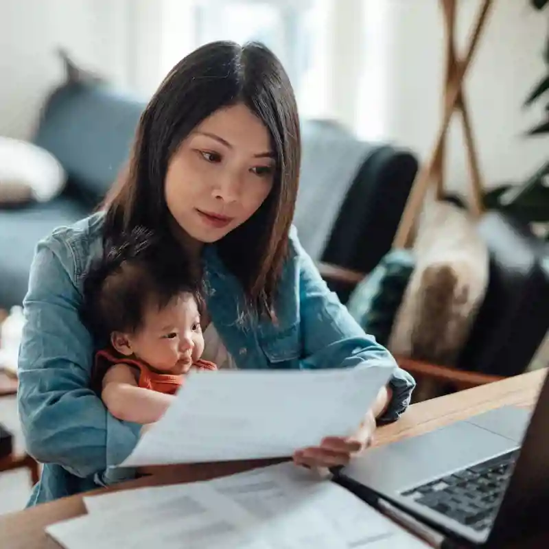 Woman looking at paper in front of laptop while holding a baby.